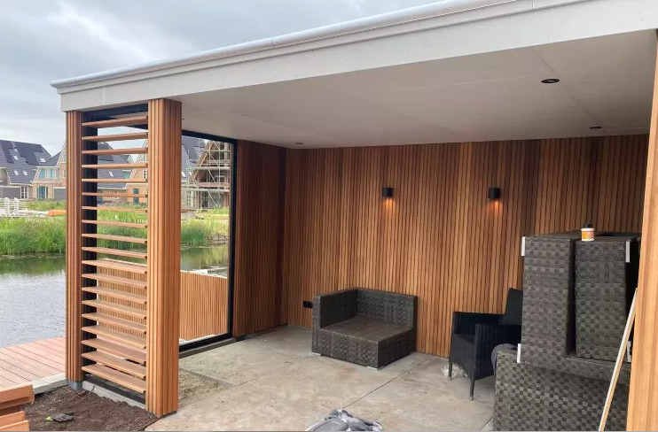 Een moderne, overdekte buitenlounge met houten lambrisering, lamellenwanden, een betonnen vloer en rieten zitmeubelen aan een kanaal.