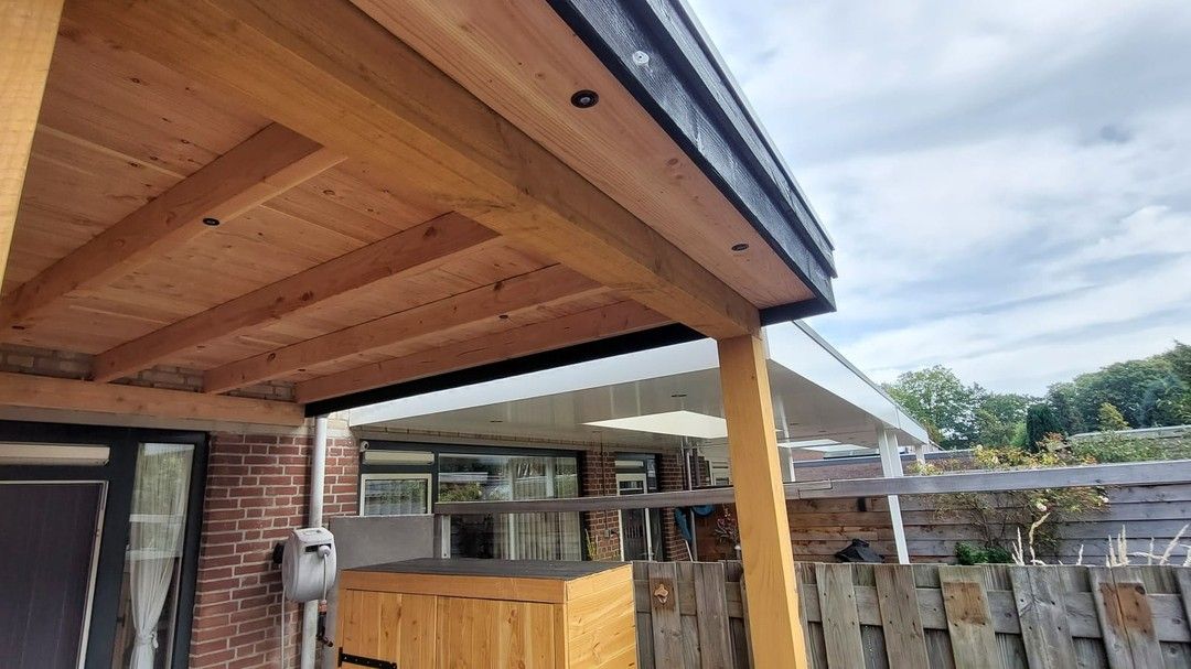 Een houten veranda met een dak en een bakstenen gebouw op de achtergrond.