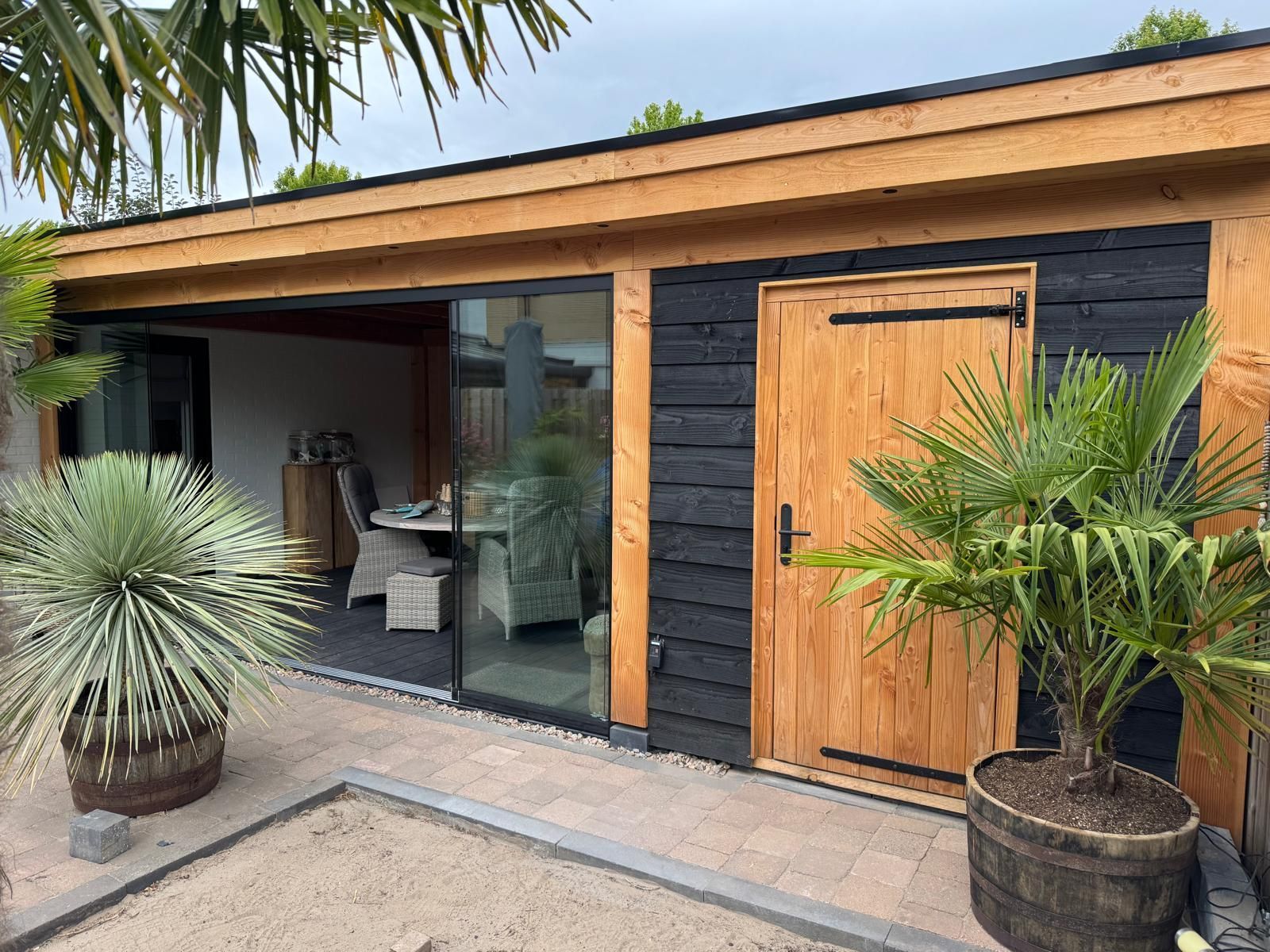 Een moderne tuinconstructie met een houten frame, zwarte gevelbekleding, een houten deur en glazen schuifwanden met uitzicht op een patio.