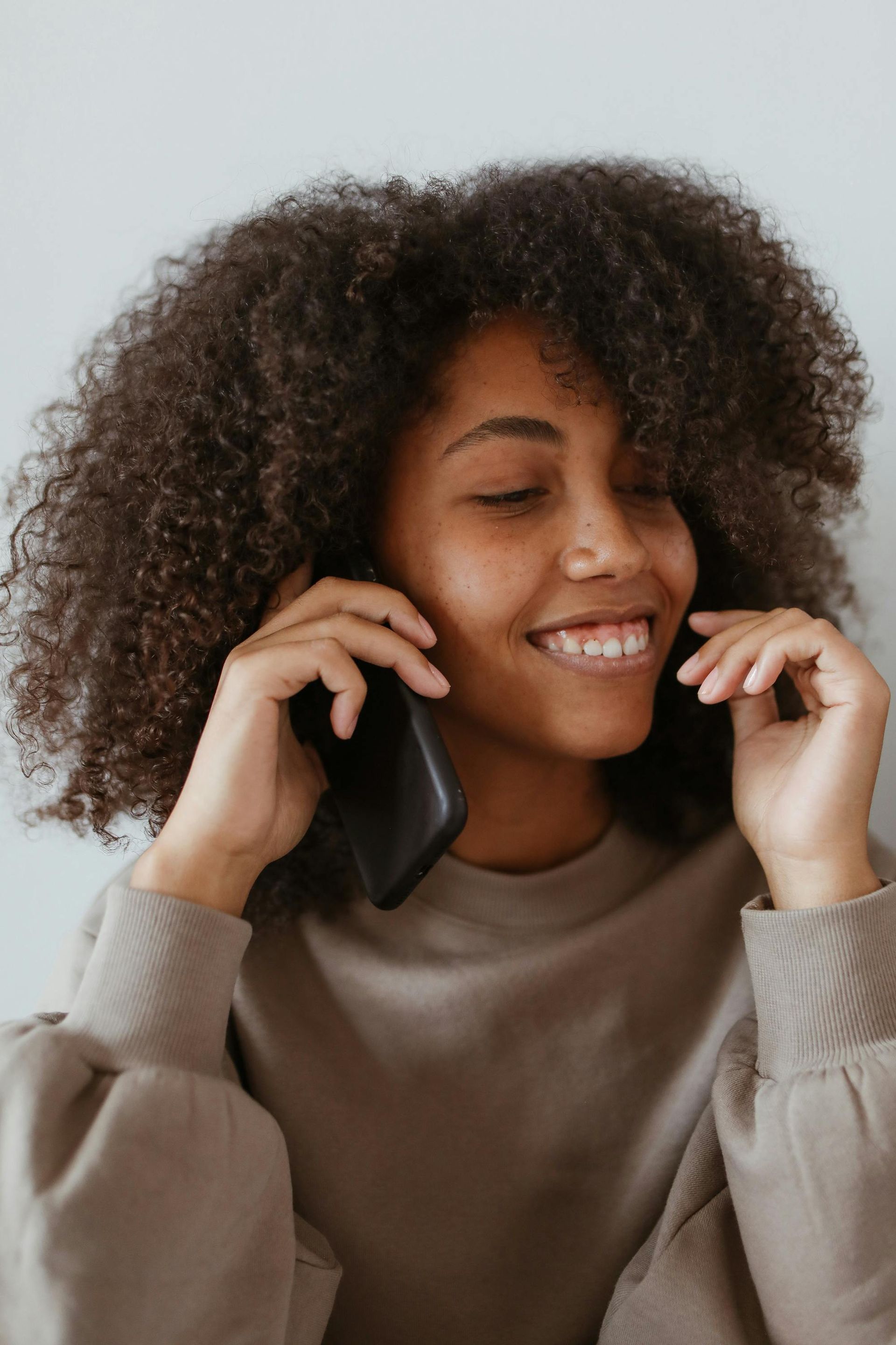 Een lachend persoon met krullend haar, gekleed in een beige sweatshirt, praat aan de telefoon op een zwarte smartphone tegen een lichtgrijze achtergrond.