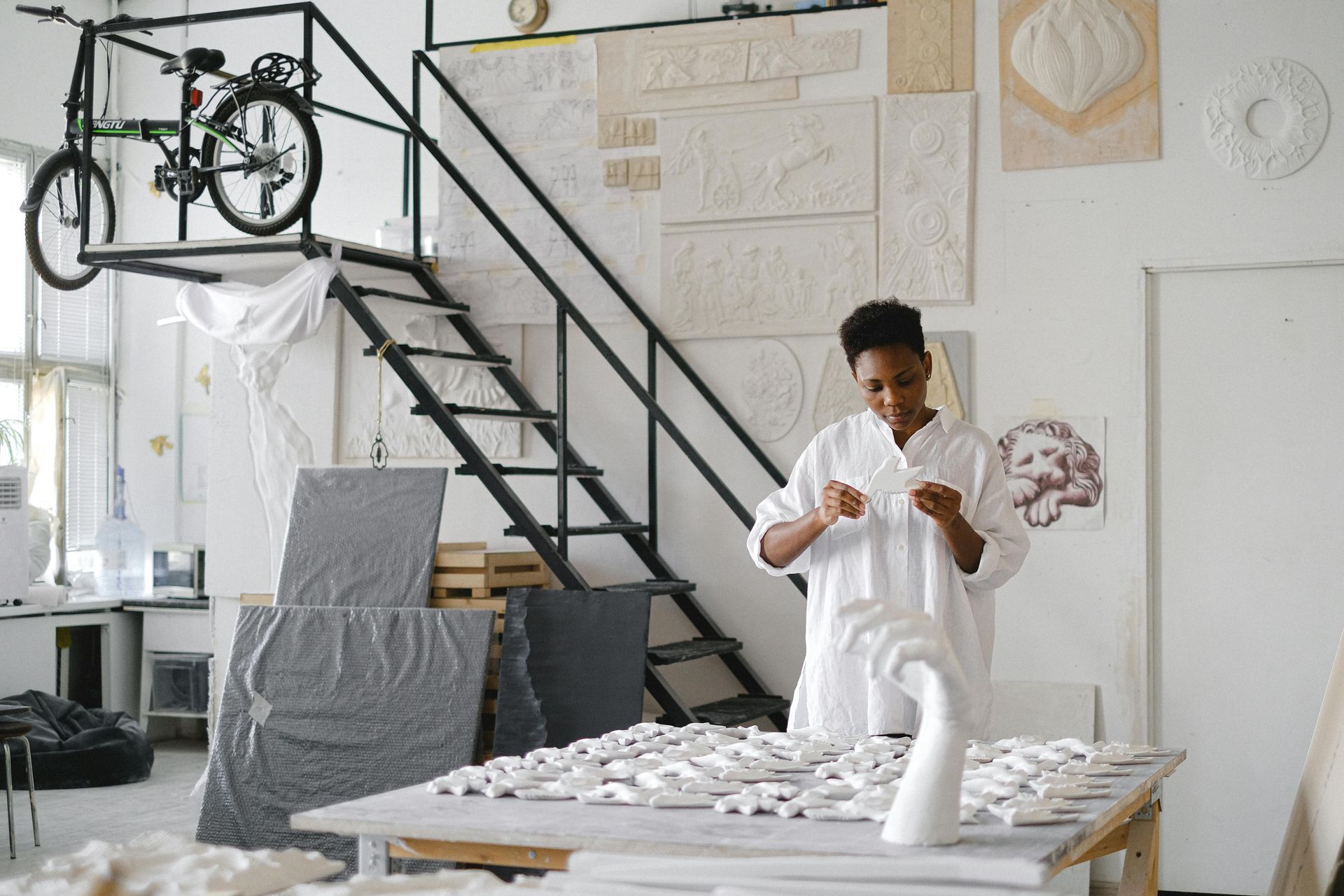 Een kunstenaar in een witte tuniek werkt in een atelier, omringd door sculpturale reliëfs, met een fiets op een verhoogd platform.