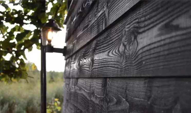 Een close-up van zwarte houten gevelbekleding met opvallende nerfpatronen, verlicht door een kleine, warme wandlamp.