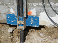 Blue metal brace holding a black pipe in a dug-up concrete wall foundation