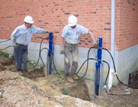 Two workers beside brick building inspect blue utility pipes in a muddy trench.