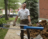 Worker standing beside a blue pipe tool, handling a pile of dirt outside a brick building.