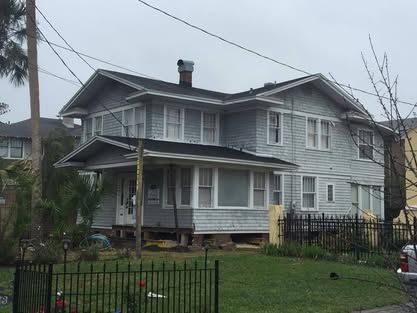 Gray two-story house with a porch and black fence on a grassy lot under an overcast sky
