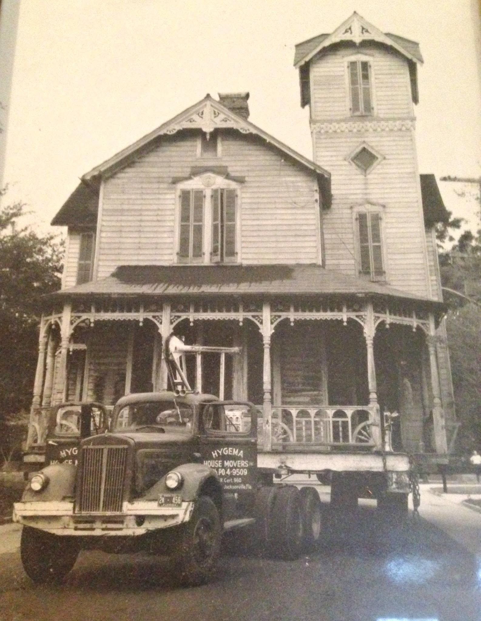 Vintage house with wraparound porch and tower, with two old cars parked in front.