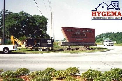 Yellow excavator and truck near a large tilted red sign by a road, with trees and a white van in the background