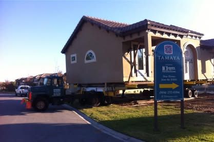 House being transported on a truck beside an AMAYA sign in a sunny roadside setting