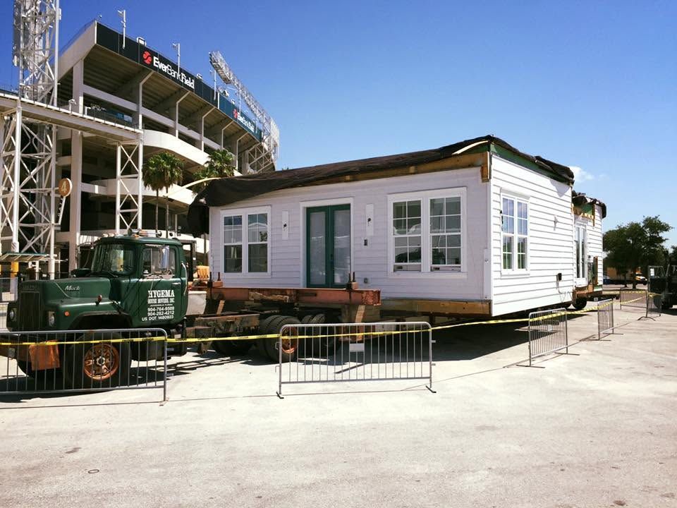 White modular house on a platform beside a stadium under clear blue sky