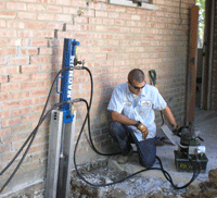 Technician drilling a concrete floor beside a brick wall with a mounted drilling rig and power unit.