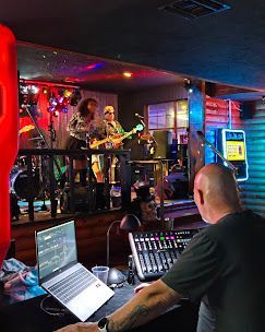 Live band performing on stage in a dim bar, seen from a sound mixer with laptop in the foreground.