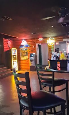 Dimly lit bar interior with high tables, stools, neon signs, and a jukebox in the background