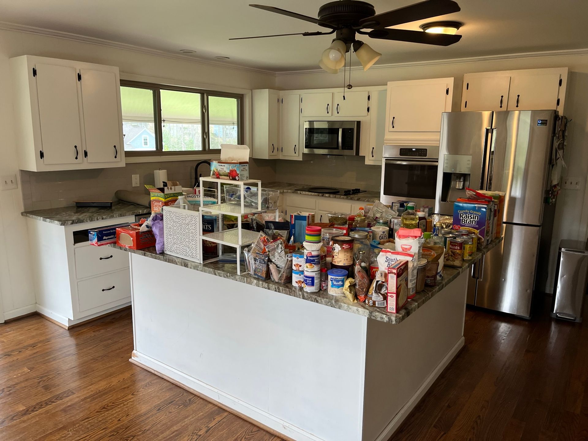 A cluttered kitchen with white cabinets, island, and stainless steel fridge overflowing with groceries.