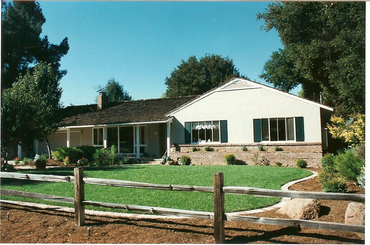 Ranch-style house with beige siding, brown roof, green lawn, wooden fence, and blue sky.