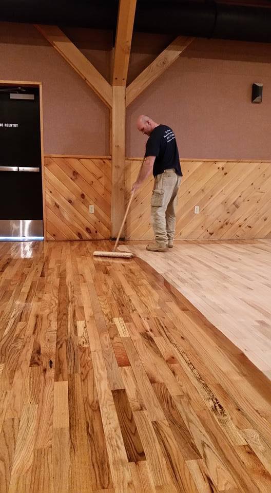 Man applying finish to a hardwood floor in a room with wood paneling and exposed beams.
