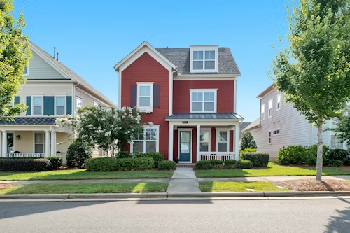 Red two-story house with white trim and blue front door, flanked by green trees on a sunny day.
