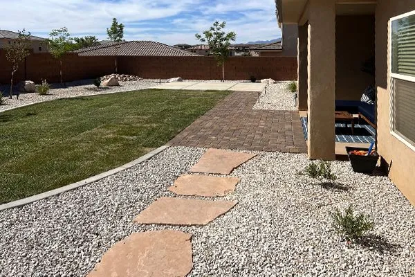 Backyard with flagstone path, gravel, grass, and a covered patio under a sunny sky.