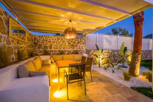 Warm outdoor patio with beige seating, wooden table, stone wall, and glowing string lights at dusk