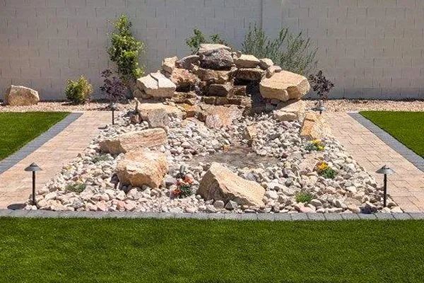 Water feature with stacked rocks, waterfall, and stone path in a backyard with green grass and white wall.
