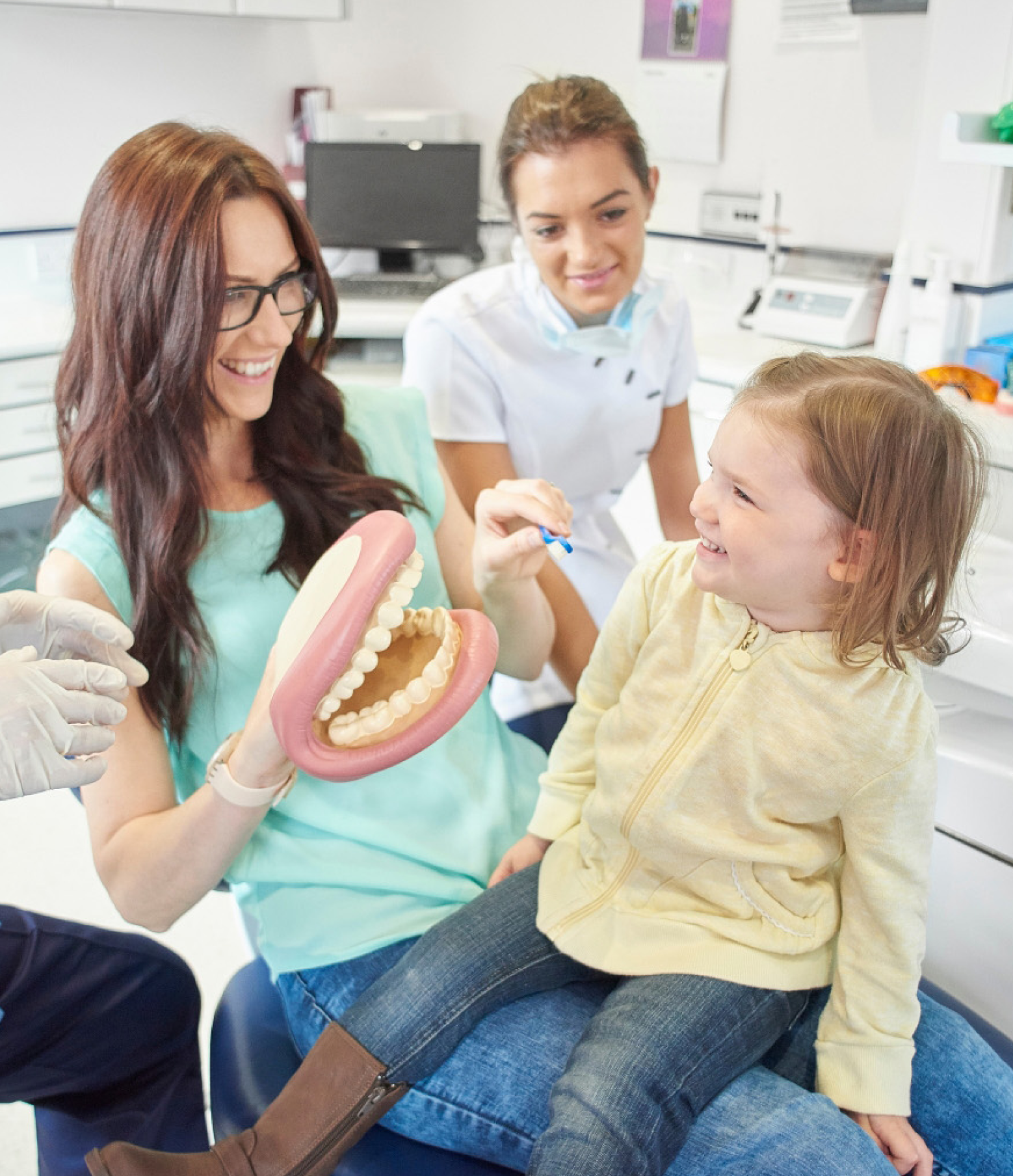 Woman showing a child a dental model; dentist assists. Bright dental office setting.
