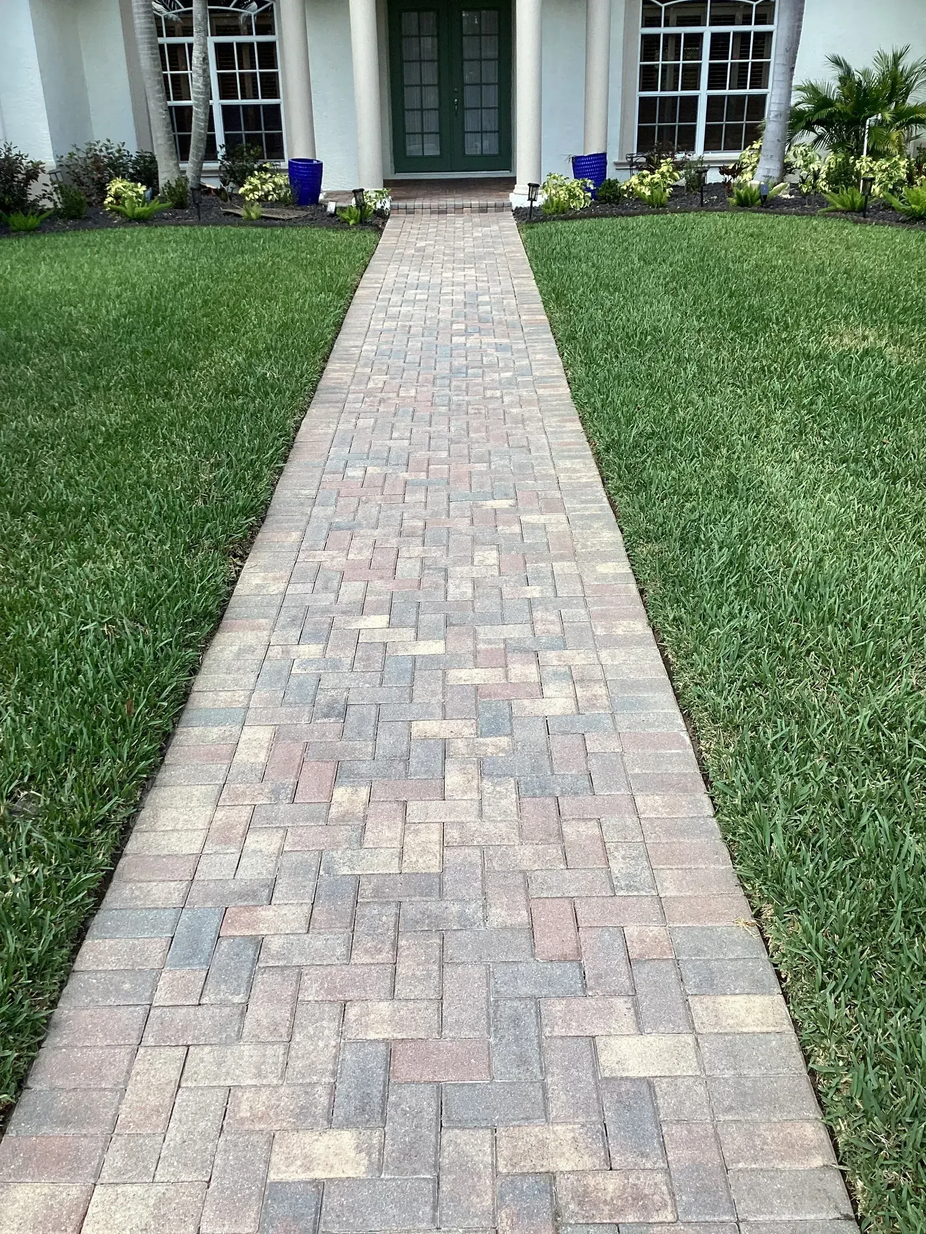Brick walkway leading to a front door, bordered by green lawn and potted flowers.