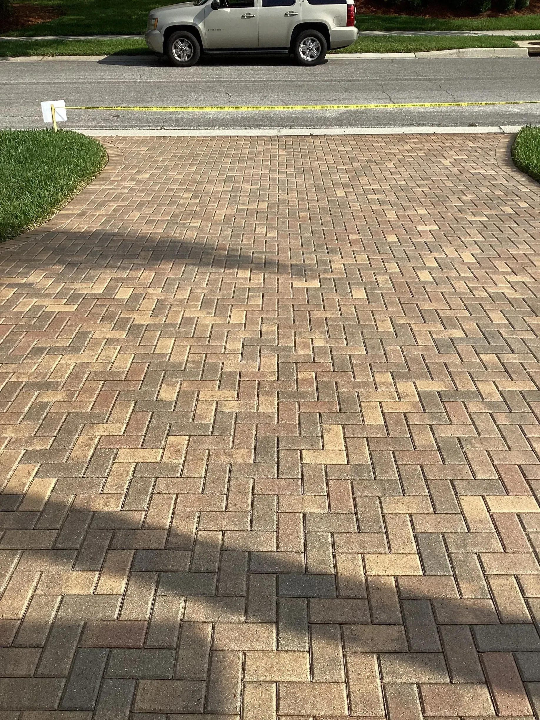 Brick driveway leading to a street, with a parked silver SUV and a grassy curb beyond.