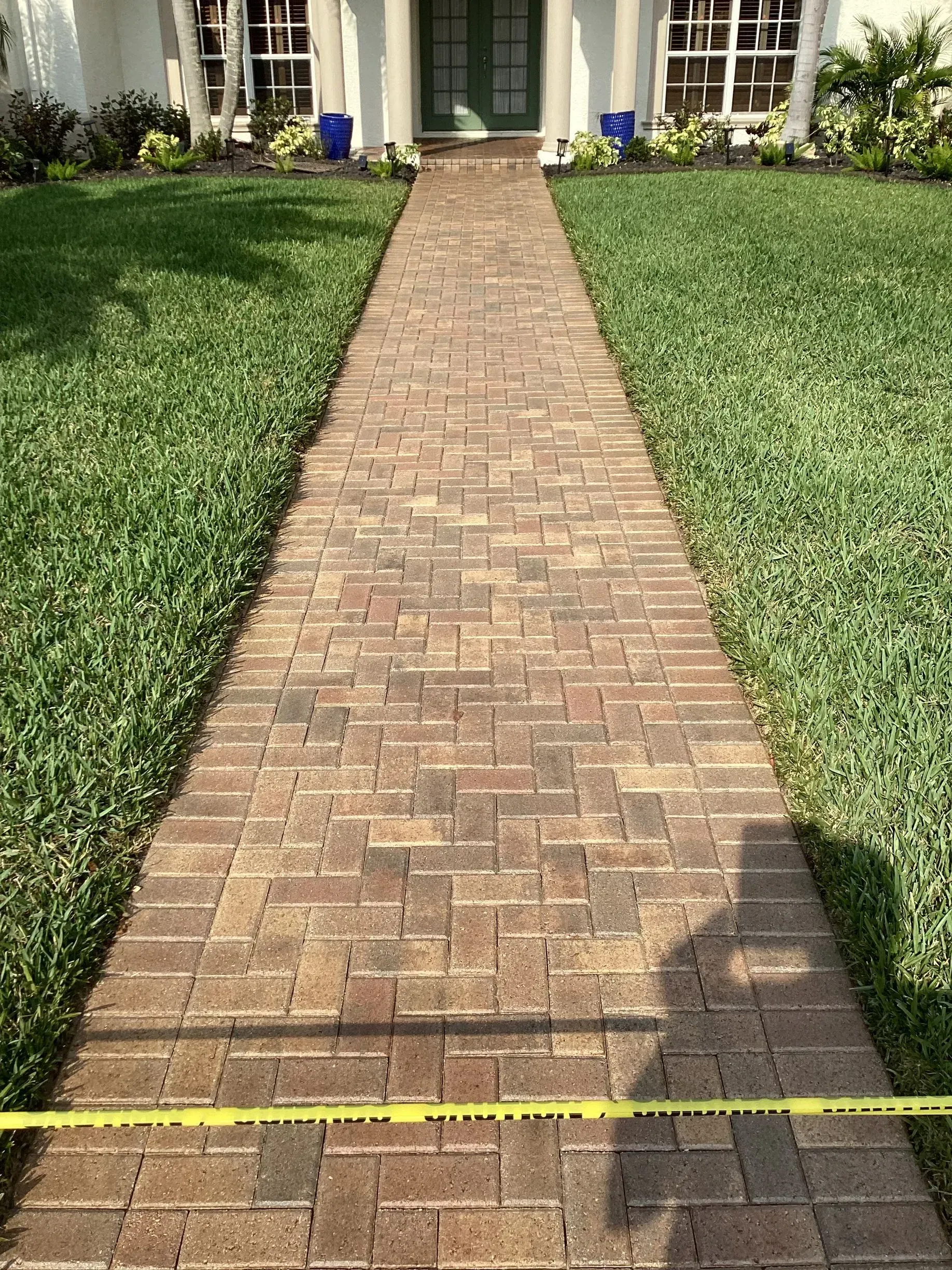 Brick walkway leading to a house, bordered by green grass and marked with a yellow tape measure.