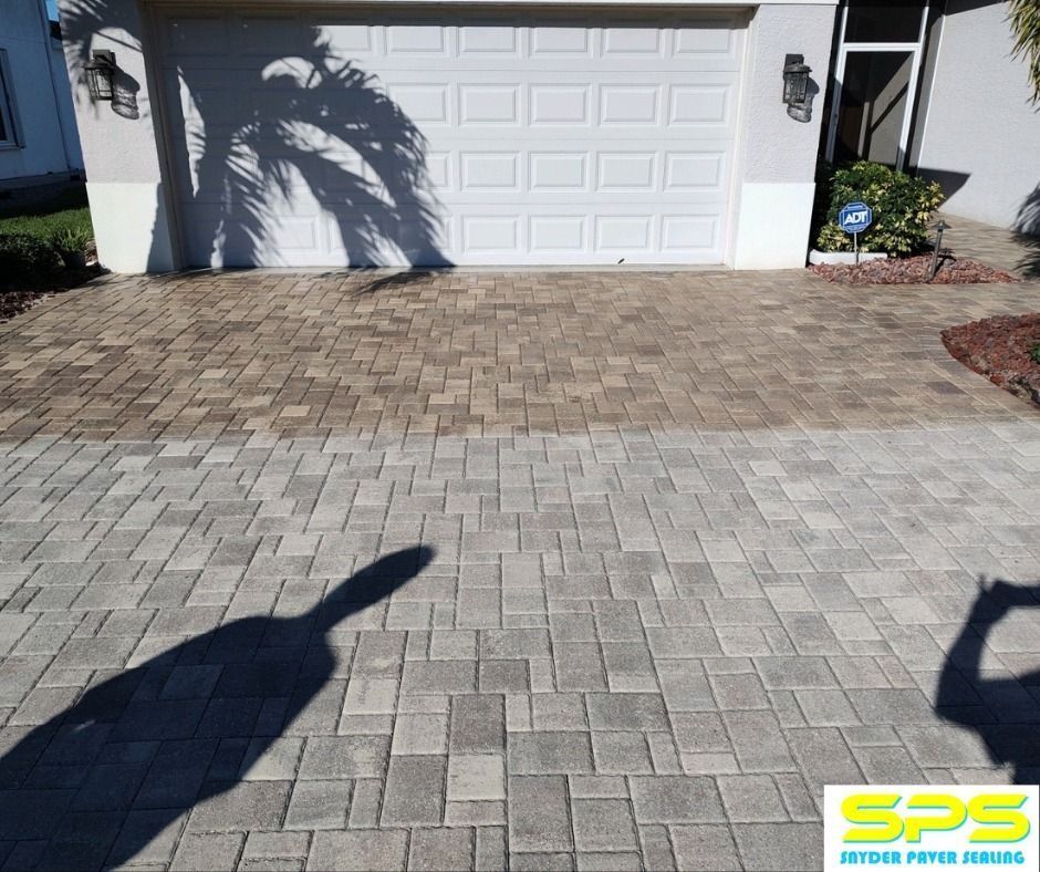 Paver driveway in front of a white garage, with dark shadows and a small landscaping bed on the right.