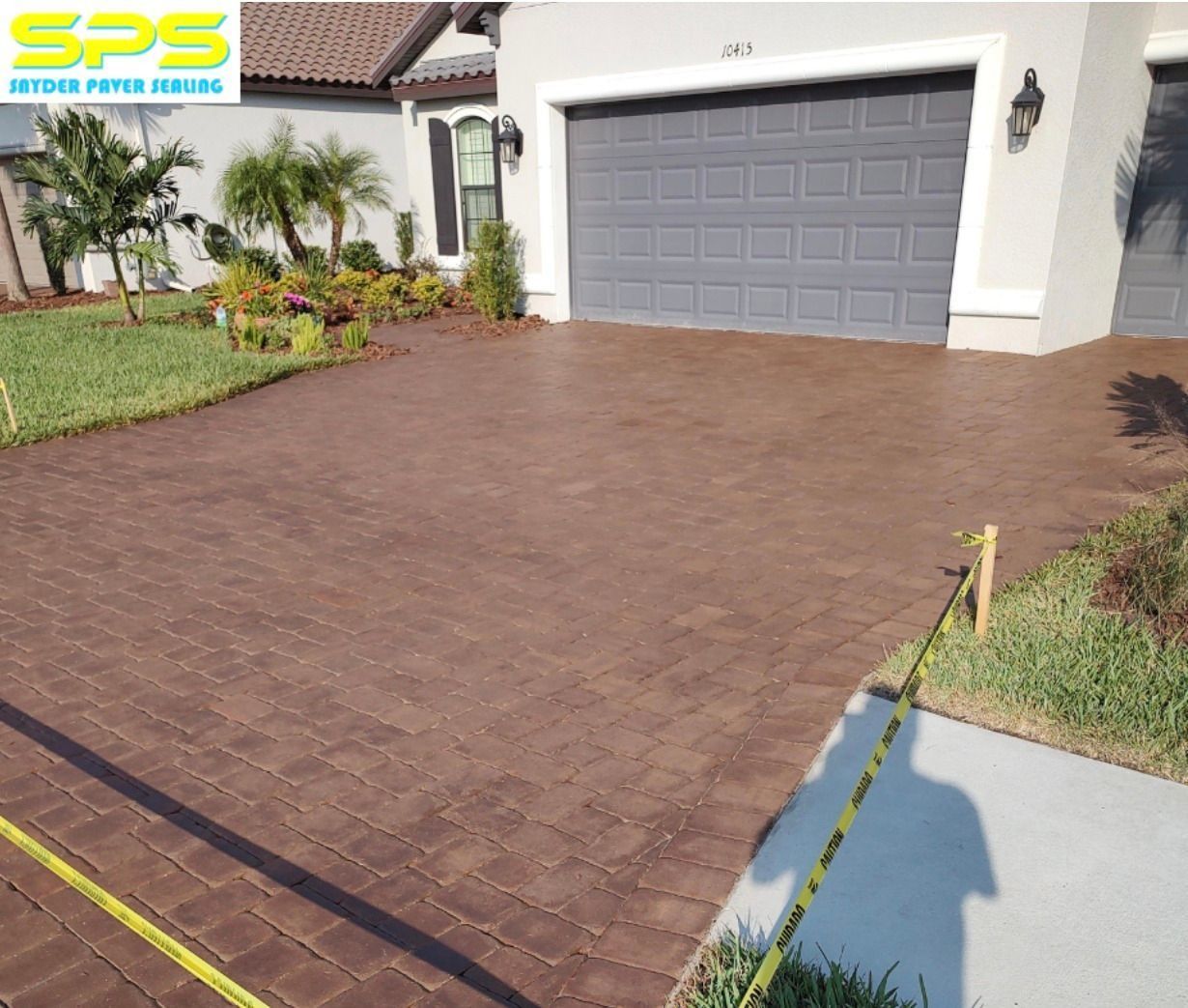 Brick driveway leading to a gray garage door beside a landscaped front yard with palm trees.
