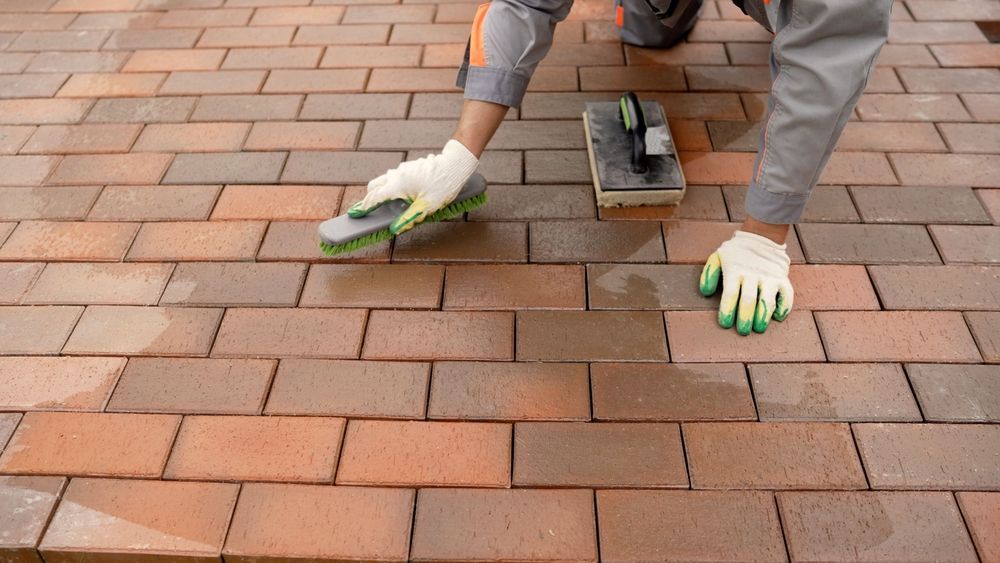 Worker laying red and tan brick pavers on a patio with gloves and a leveling tool