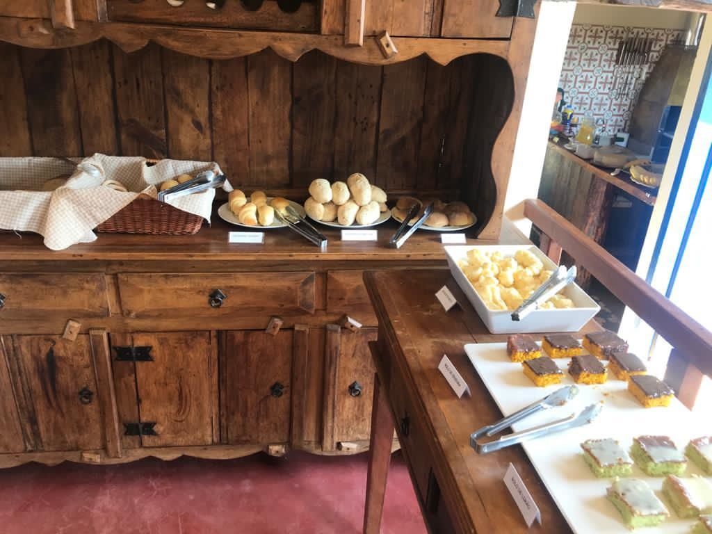 Mesa de buffet de madeira com diversos produtos de panificação: pãezinhos, pão de queijo e mini bolos.
