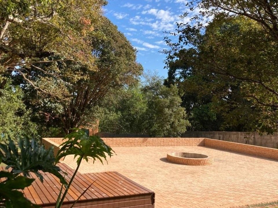 Pátio de tijolos com lareira, rodeado por árvores sob um céu azul.