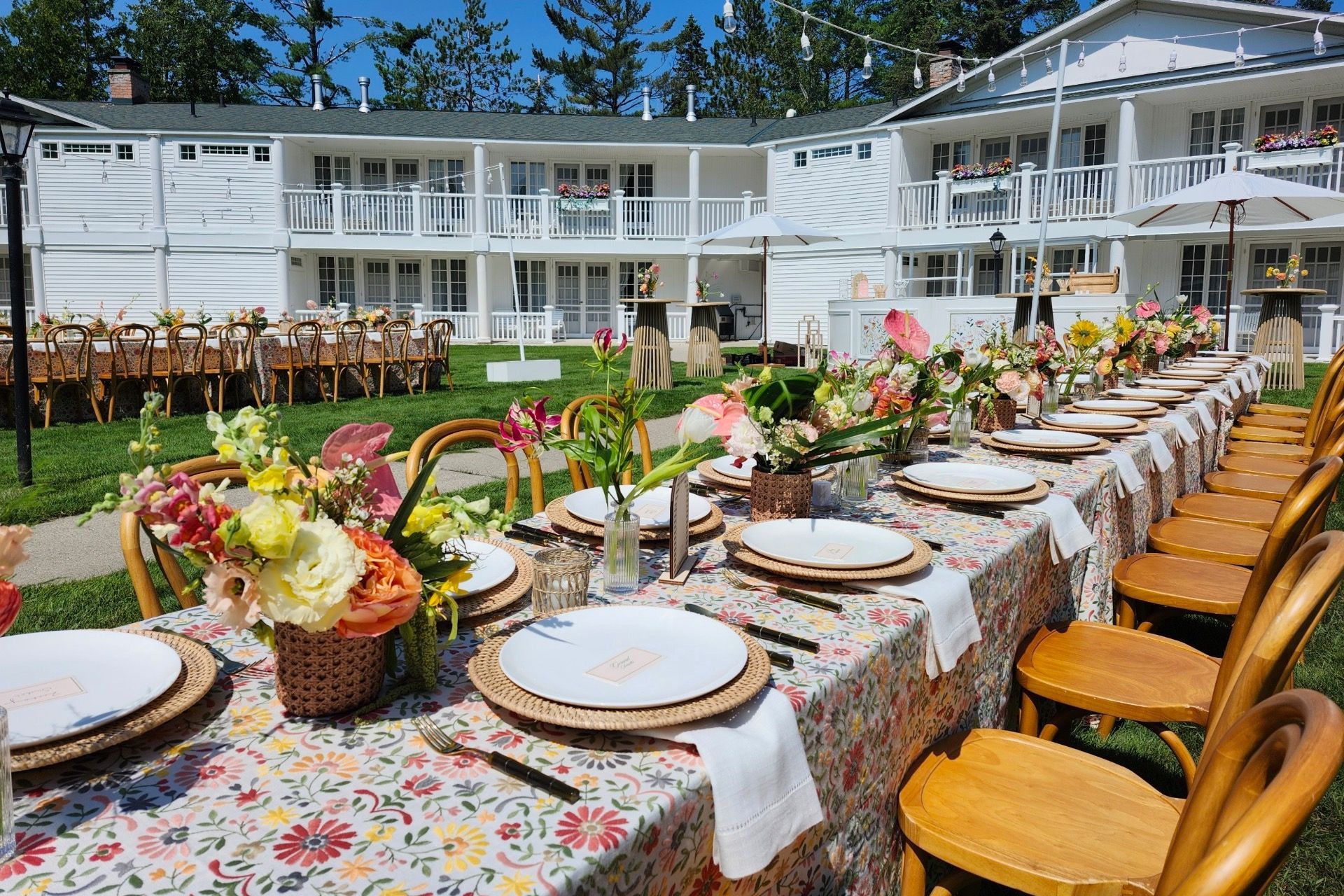 Long outdoor dining table set with plates and floral centerpieces beside a white house and lawn