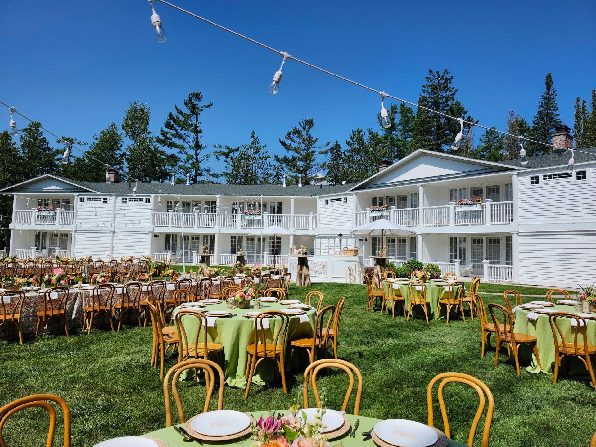 Outdoor dining setup with tables and chairs on a lawn in front of a white building under a blue sky