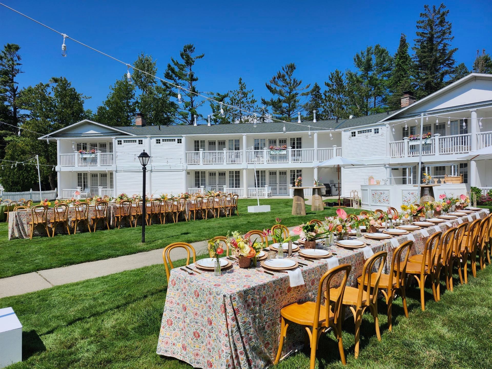 Outdoor wedding reception with long floral tables and wooden chairs in front of a white lodge under a blue sky