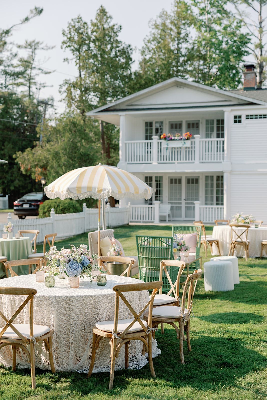 Outdoor garden party setup with round table, white and gold chairs, and striped umbrella in front of a white house
