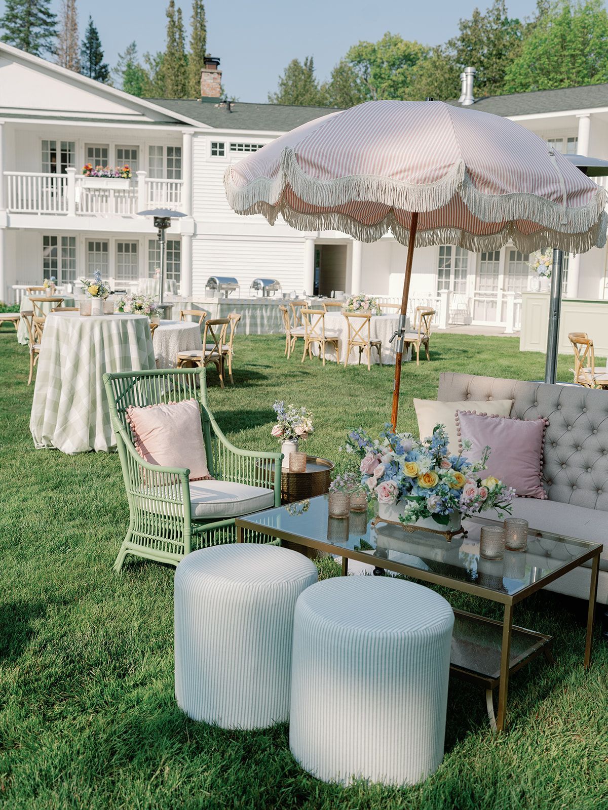 Elegant outdoor lounge with pastel chairs, glass tables, and a pink umbrella on a lawn beside a white house.