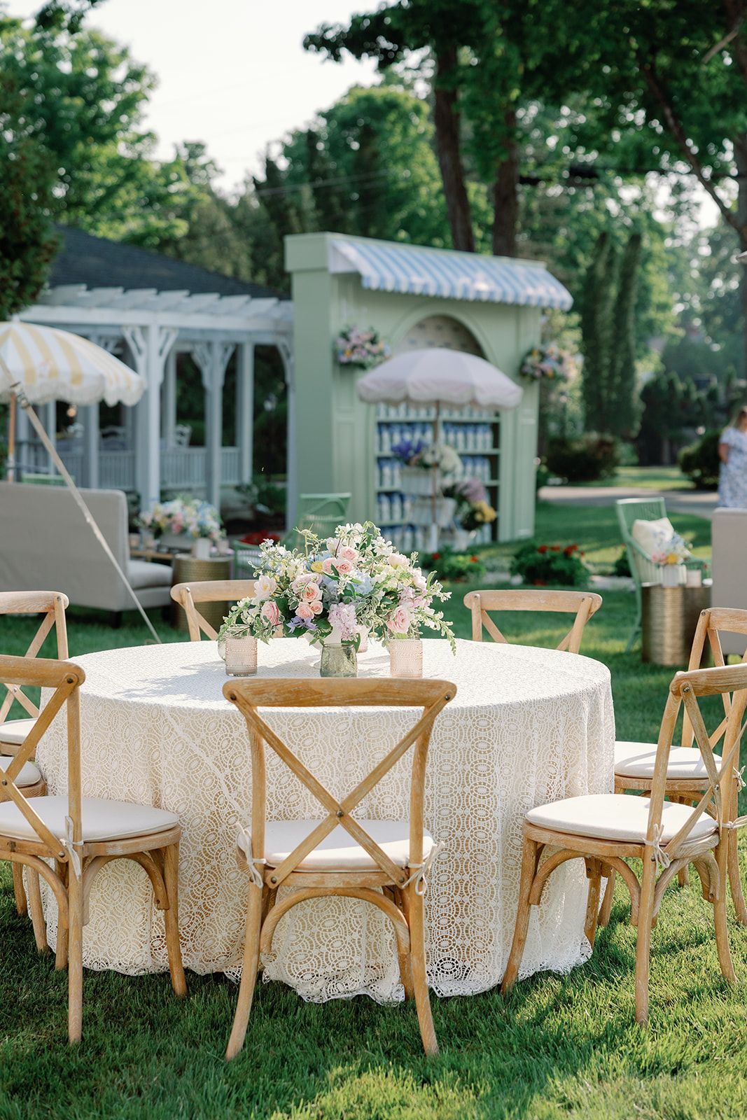 Outdoor event setup with round lace-covered tables, wooden chairs, and floral centerpieces on a grassy lawn