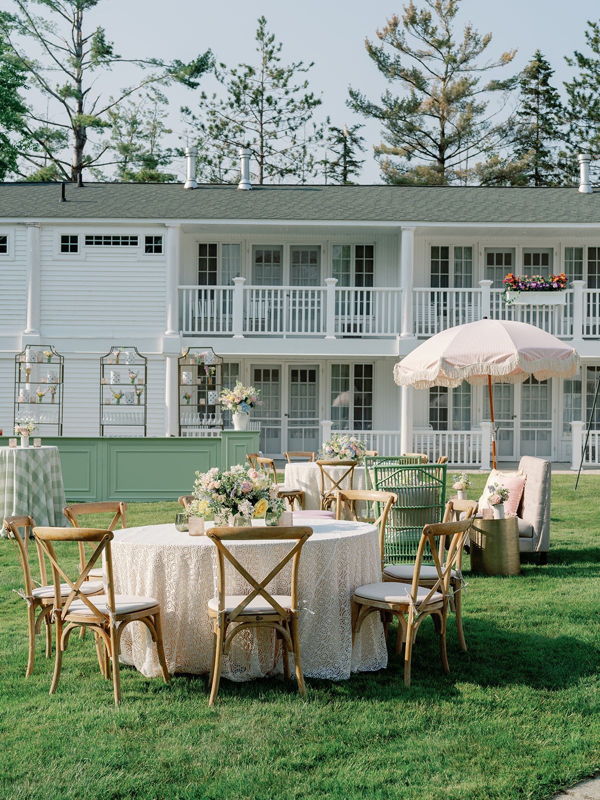 Outdoor dining setup on a lawn beside a white two-story house, with tables, chairs, and a pink umbrella.