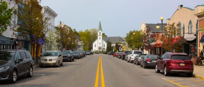 Small-town main street with parked cars, storefronts, and a white church at the end of the road.