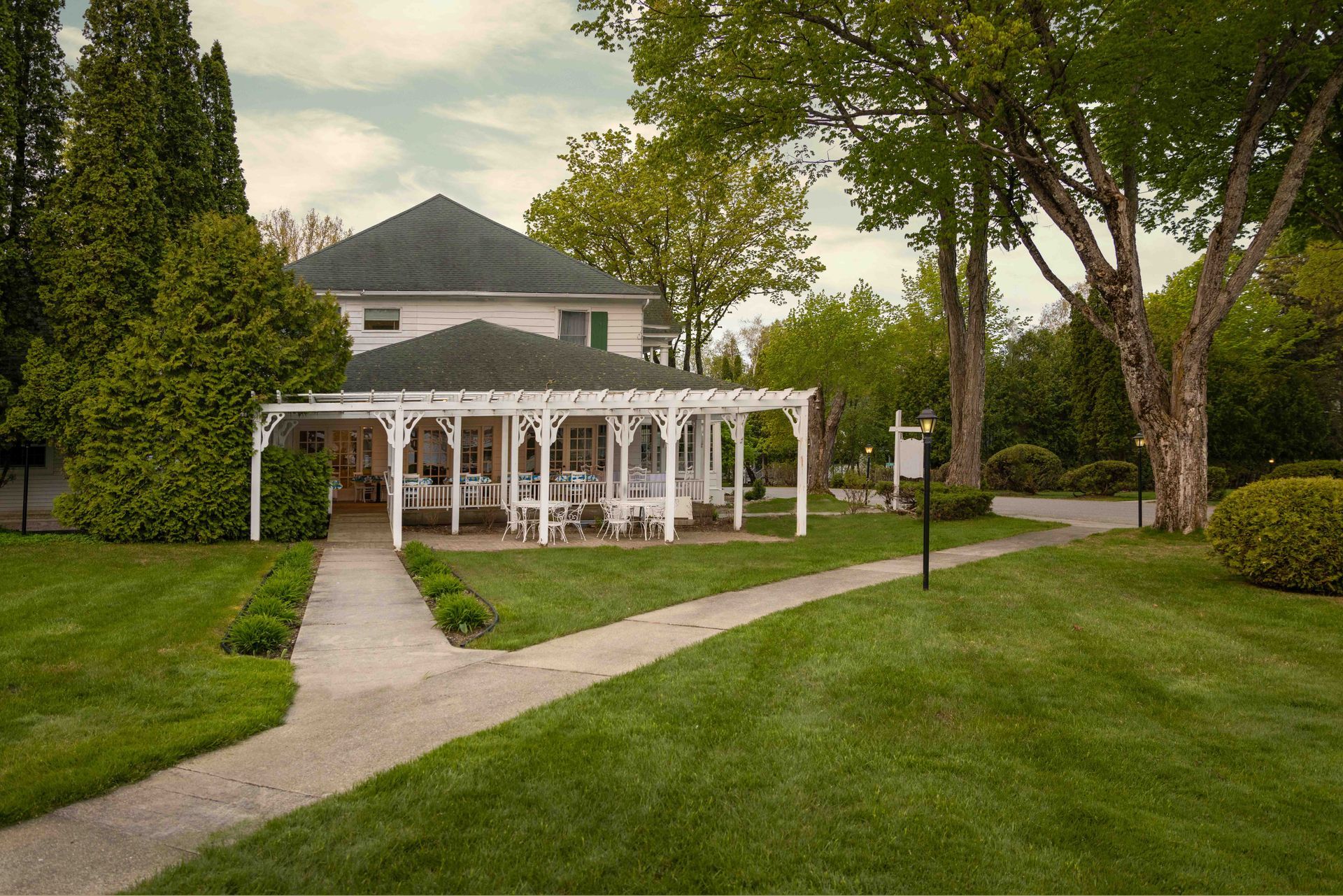 White house with wraparound porch beside a winding path, surrounded by green lawn and trees