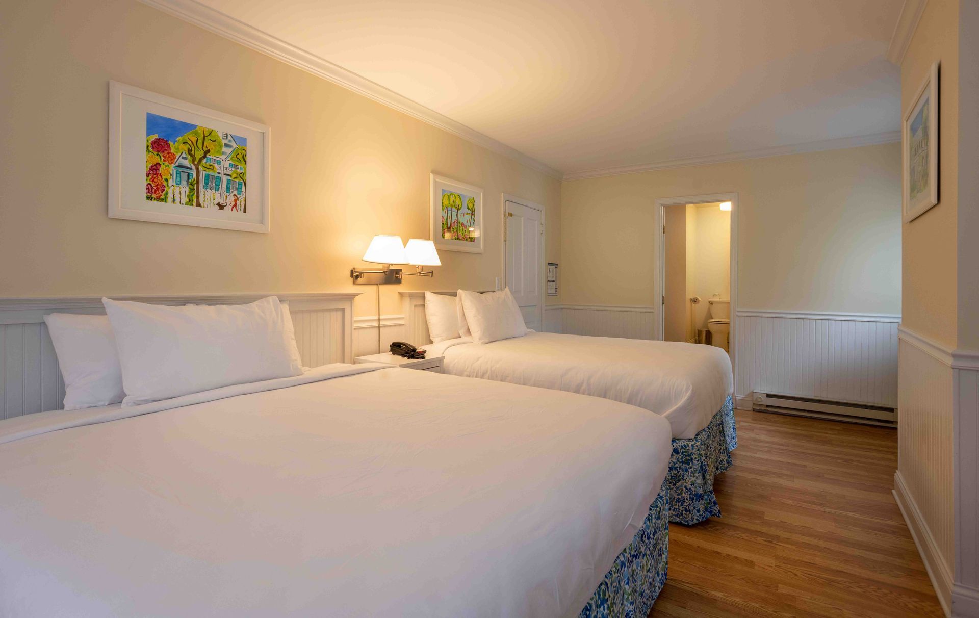 Two neatly made twin beds in a bright beige hotel room with artwork and a bedside lamp