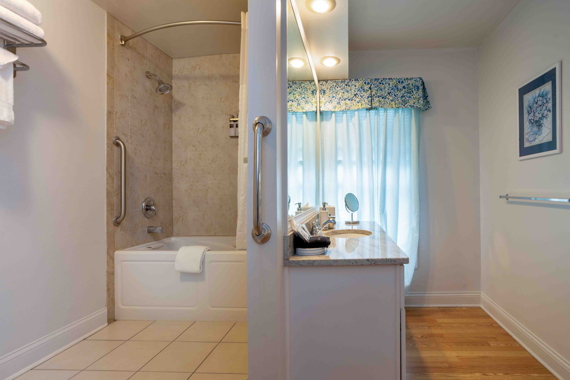 Modern bathroom with beige tile, white tub and shower, beside a bright kitchen or hallway area with curtains
