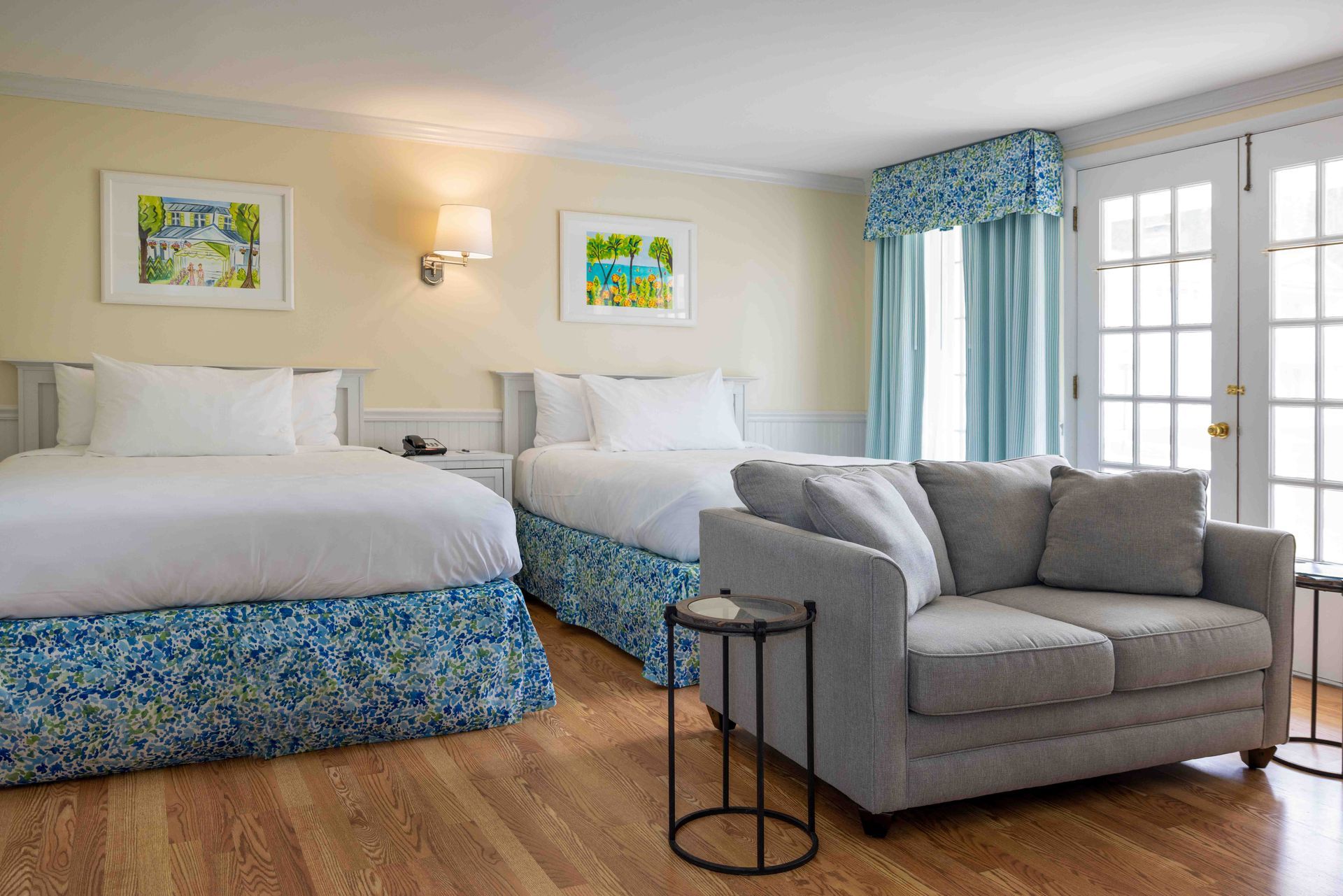 Hotel room with two beds, gray sofa, blue accents, and French doors to a balcony