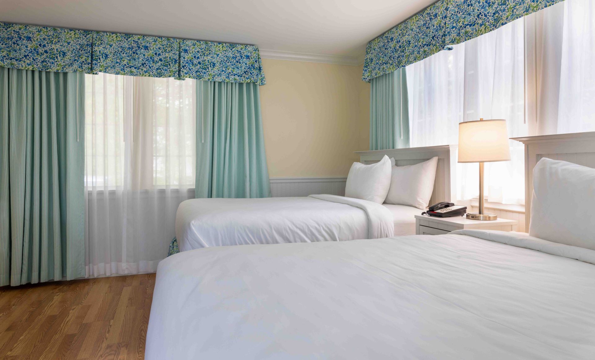Hotel room with two white beds, blue curtains, and a bedside lamp by a large window
