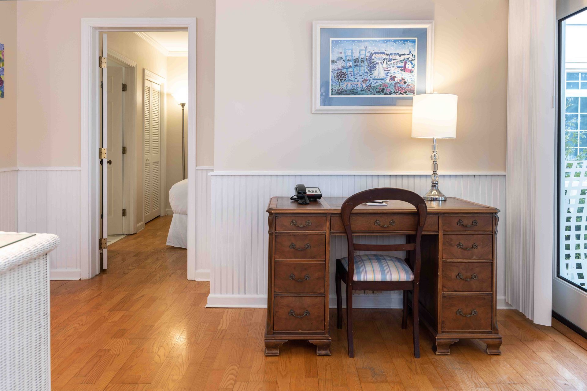 Home office with wooden desk, lamp, framed art, and hardwood floors in a bright room