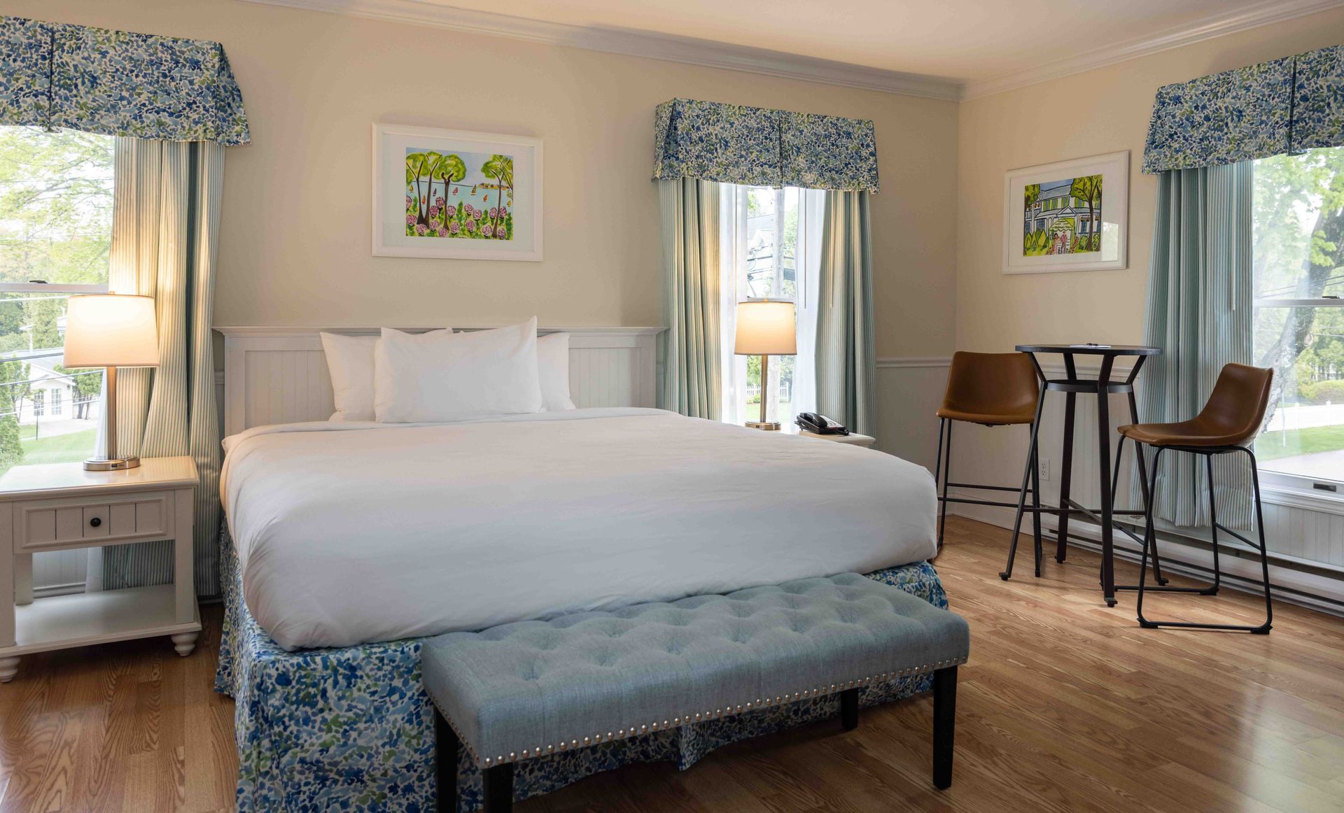Bright bedroom with a large bed, blue bench, two windows, and a small table with stools.
