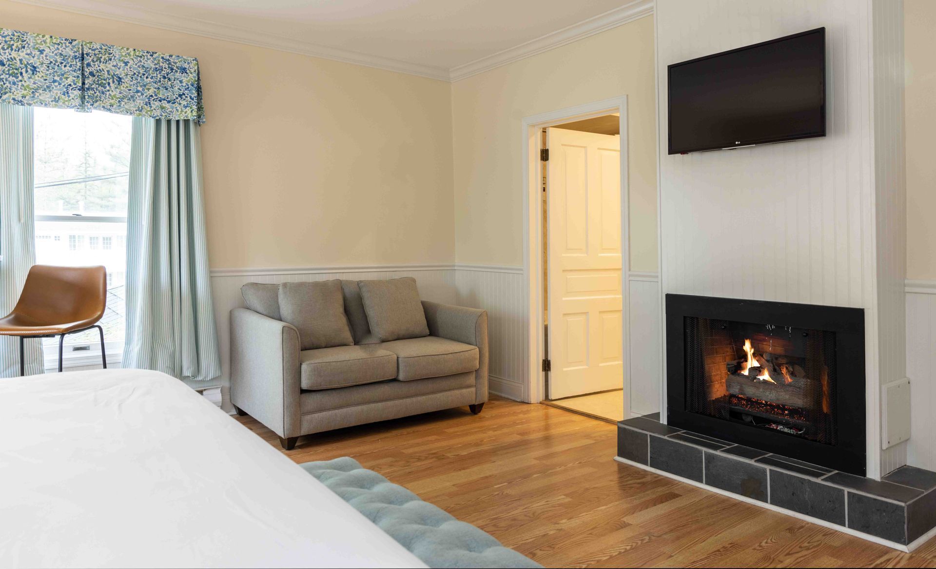 Bright bedroom with bed, gray sofa, wall TV, and a fireplace on a light wood floor.