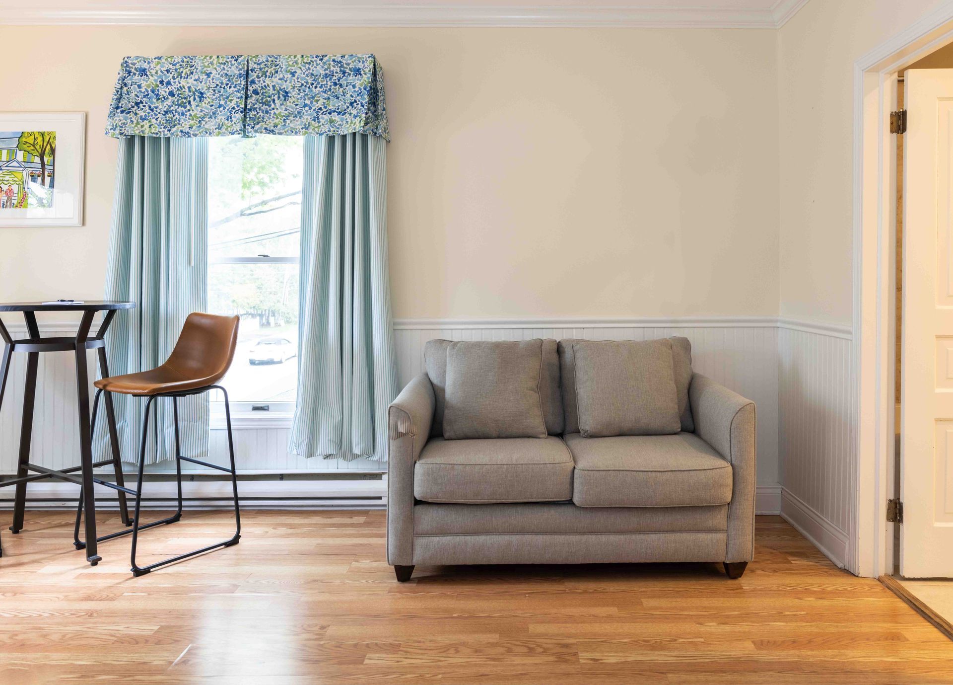 Bright living room with a gray loveseat, tall window with blue curtains, and a wooden floor.